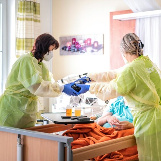 Zwei Pflegerinnen in Schutzkleidung arbeiten am Pflegebett eines Patienten