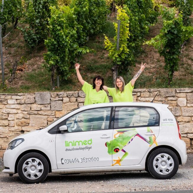 Zwei Pflegerinnen winken hinter einem Auto der häuslichen Krankenpflege